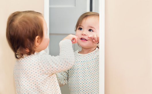 Happy toddler girl looking at herself in the mirror in her house
