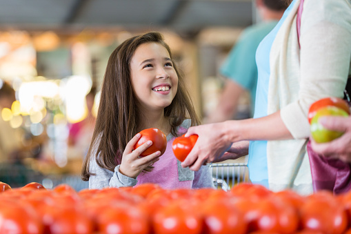 Elementary age Hispanic little girl is smiling while shopping for fresh produce at market or local grocery store with her mother. Child is holding a ripe red tomato in produce section and smiling while looking up at her mom.