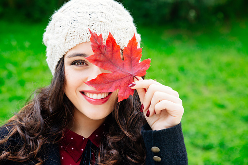 Beautiful young woman with woolen cap covering face with a red leaf while smiling against a green background. Fresh skin and healthy smile.