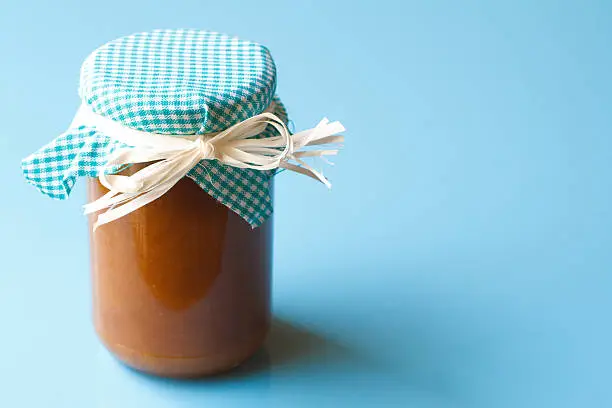 A vintage-style homemade jar of jam (or marmalade or fruit preserves) on a pale blue background with plenty of copy space. The jar is covered with blue and white checked gingham fabric tied on with a straw bow.