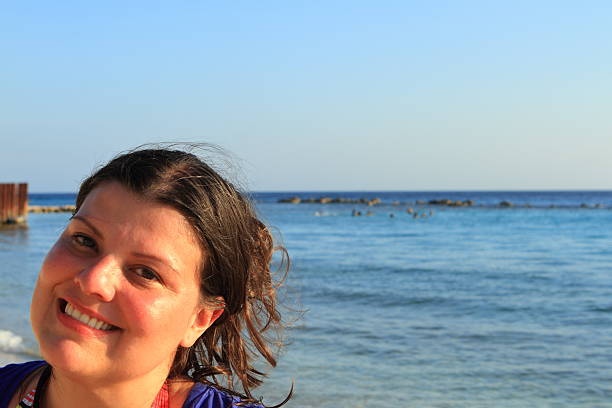 Lady smiling poses for a close-up shot. She is at a beach with blue water.