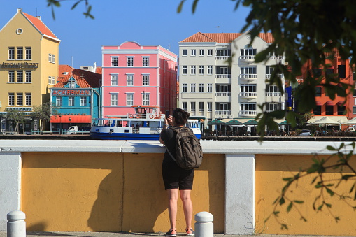 Lady is taking photograph of the St. Anna Bay in Curacao. She is using her smartphone and has a backpack, She is wearing black short pants.