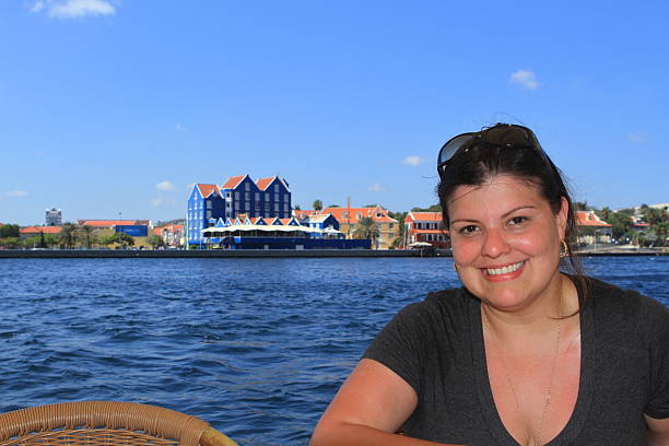 Lady is smiling while posing for a portrait next to the St. Anna Bay in Curacao. Fill flash was used lo illuminate the face of the model and have some good color for the background that identifies crearly the location. She is wearing sunglasses on her head.