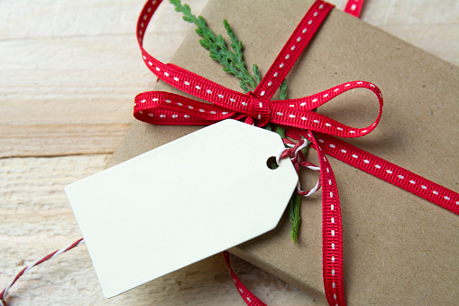 Gift box, wrapped in recycled paper, red bow and empty tag on wood background