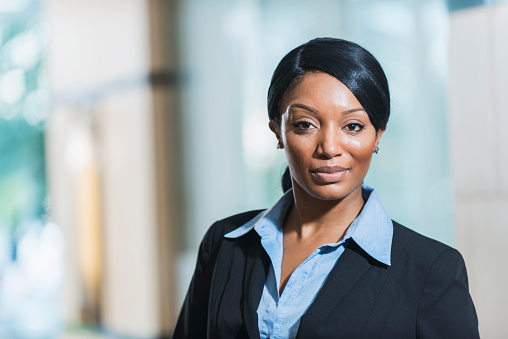 Portrait of an African American businesswoman standing in an office lobby, near a window, looking at the camera with a serious, confident expression.  She has straight, long, black hair hair back in a ponytail.  She is wearing a black suit.
