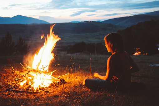 Vintage toned, low key image of a young boy, making a campfire in the nature as the sun sets down.