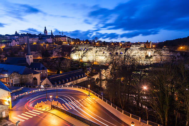 Picture of the old city of Luxembourg, and the old suburb Grund in the below. Picture taken just after sunset, with cars leaving yellow and red light trails on the road. Nice clouds and blue sky.