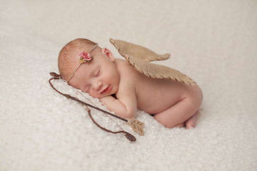 Portrait of a smiling, red headed, 2 week old, newborn baby girl. She is wearing a Cupid costume with angel wings, bow and arrow and is sleeping on a cream colored blanket.