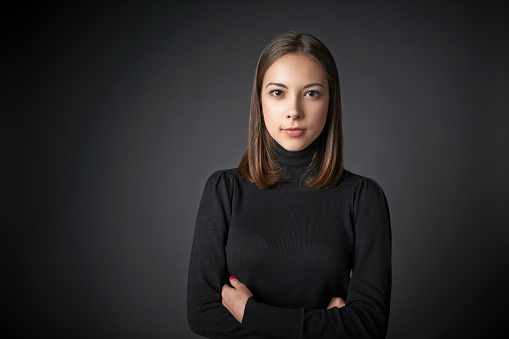 Closeup portrait of young female in black pullover with  folded hands with a speculative look on her face looking at the camera, over dark studio background