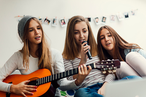 Teenage Girls Having fun together at slumber party. Sitting on couch and playing guitar and singing karaoke. Looking song lyrics on laptop. Shot with Canon EOS 5Ds 50mp. Selective focus to girl with microphone.