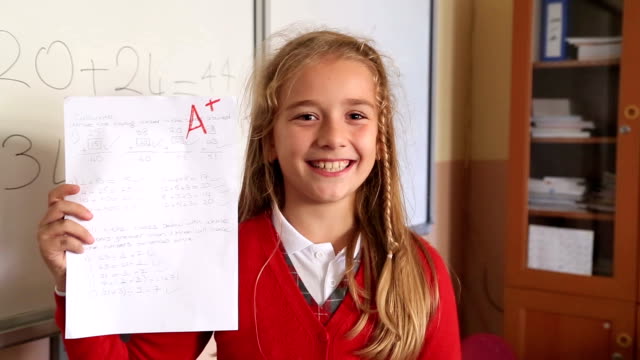 Happy cute little girl showing exam paper with a plus in the classroom