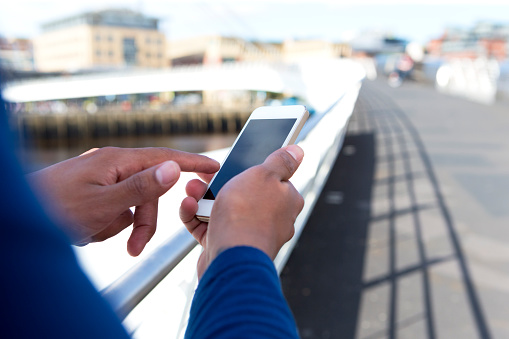 A pair of male hands are holding a smartphone and texting whilst on the go. There is an urban scene in the background and a winding bridge.