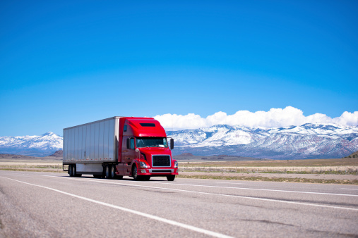 Large beautiful modern classic-modern red truck with a high cab and trailer on a flat stretch of highway on a background of snow-capped mountain ranges, drowning in the clouds and clear blue sky