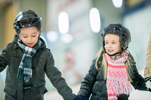 A brother and sister are ice skating together at in indoor skating rink. They are holding each others hand while skating.