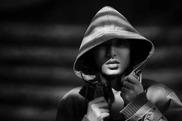 Photo of Young woman in hoodie jacket under dark shadows of blinds.