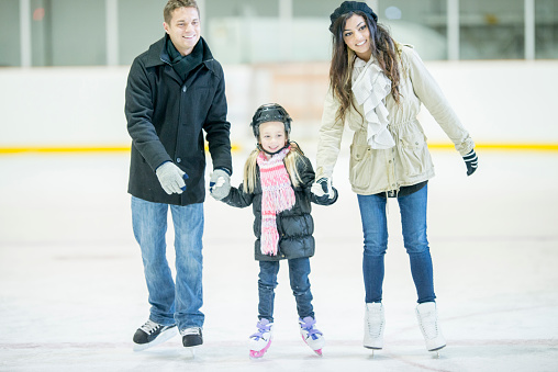 A cute little girl is learning how to ice skate at is holding onto her mother and father. She is wearing a helmet and smiling.