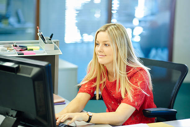 young female office worker or intern young female office worker or work experience student , sitting at her desk and looking at her computer. school receptionist stock pictures, royalty-free photos & images