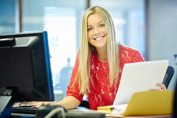 young female office worker or work experience student , sitting at her desk and looking at camera