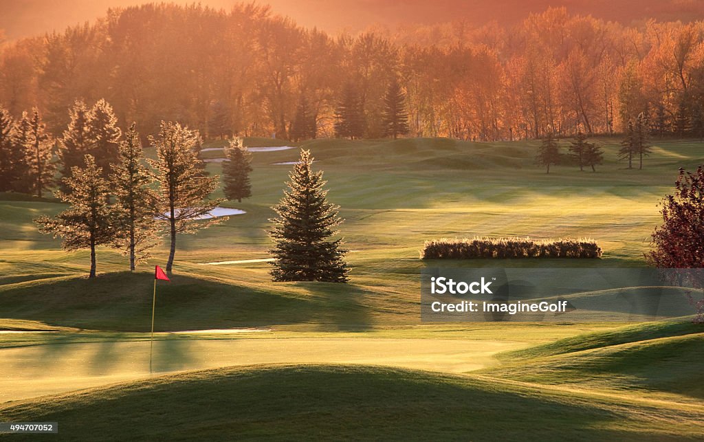 Beautiful Golf Course in Fall A stunning golf course in the fall with backlit green, red flag, and pristine shaping and fairway turf. Golf course is located in a river valley. Nobody is in the image. Image taken in peak of fall colours with aspen trees backlit on the edge of the course. Evergreen trees, aspens, and red flag highlight the scene. Golf course is situated along the banks of a large river. Autumn golf scenic at world class golf facility. 2015 Stock Photo