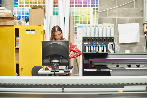A young woman is working on a digital printing machine . she is at the computer terminal loading in the next print job .