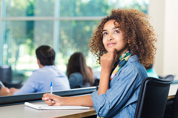 Beautiful teenage African American high school student is looking up with her hand on her chin. She is taking a test in modern high school classroom. Girl has curly hair and is wearing trendy clothing. Classmates are seated in background.