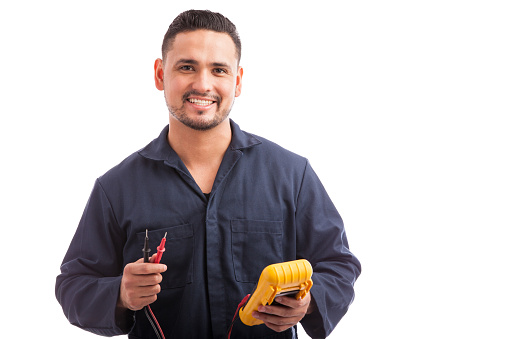 Portrait of a young Hispanic electrician wearing overalls using a multimeter and smiling on a white background