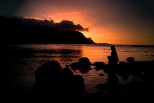 Woman sits alone on a beach at Princeville, Kauai and watches the sun set behind a bank of clouds and the Kauai mountains.  Sunset tints the sky and waters a brilliant orange and pink.