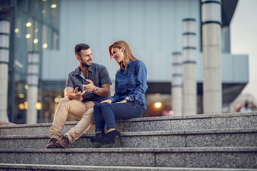 Smiling business couple sitting on stairs outdoors and talking about some text message businessman has received.
