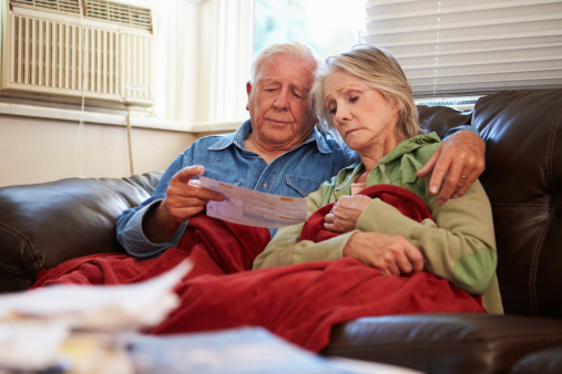 Worried Senior Couple Sitting On Sofa Looking At Bills Under A Blanket