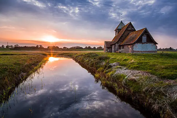 Old abandoned Norman church by the side of a river in Kent, UK. The cloudscape is reflected in the still water of the river as the glowing sunset dips below the horizon. Slight hint of lens flare and the image was shot into the sun. A peaceful, tranquil and spiritual rural scene. Horizontal colour image with plenty of room for copy space.