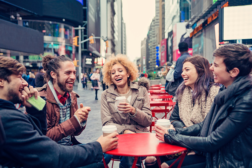 Bunch of friends sitting and laughing, drinking coffee at that public space with red tables and chairs in Times Square, New York. Three girls and two boys, hip style, mixed race in their twenties. Focus on the girl in the middle. People and buildings in the background. Horizontal shot outdoors on a rainy day.