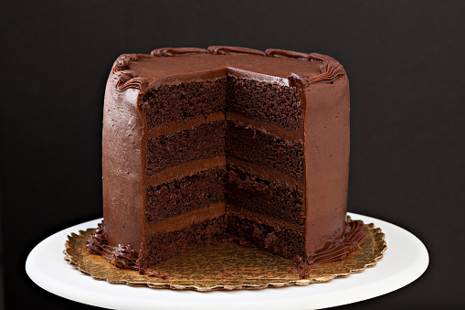 A close up, horizontal, isolated on black photograph of a multi layered chocolate fudge cake with a slice missing.