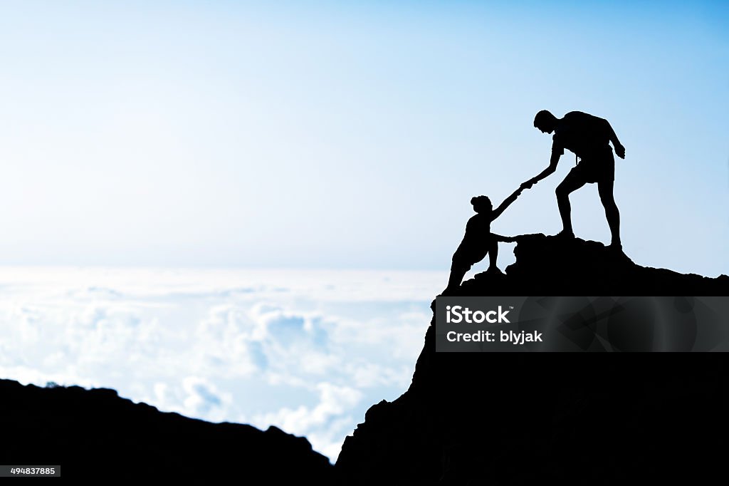 Man and woman help silhouette in mountains Couple hiking help each other silhouette in mountains, sunset and clouds. Male and woman hiker helping each other on top of mountain climbing, beautiful sunset landscape. A Helping Hand Stock Photo
