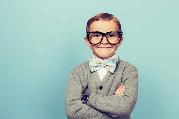 A young boy dressed as a nerd with glasses is looking in the camera with a big smile on his face. He has a bow tie on and is in front of a blue wall.