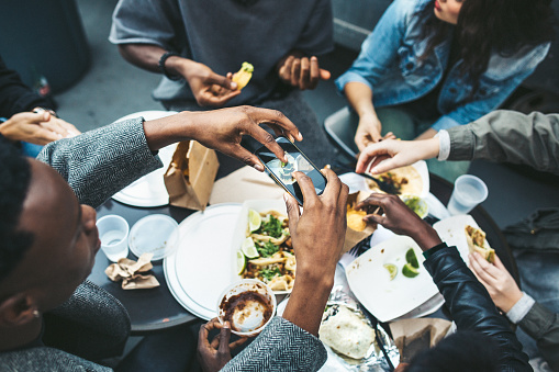 A multi-ethnic group of friends enjoy tacos and chips at a Mexican food cart in downtown New York City, New York.  They laugh and talk while eating their food, having fun sharing life and culture.  One of them takes a picture of their food with his smartphone to share on his social networks.  Horizontal image.