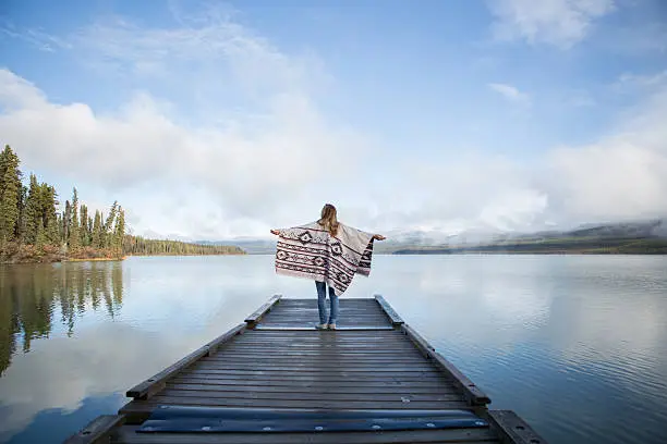 Young woman stands on wooden pier in Canada, arms outstretched for freedom and positive emotions. She is wearing a poncho. Morning sunlight passing through the mist.