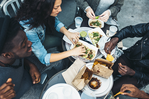 An overhead view of a multi-ethnic group of friends enjoying tacos and chips at a Mexican food cart in downtown New York City, New York.  They laugh and talk while eating their food, having fun sharing life and culture.  Horizontal image.