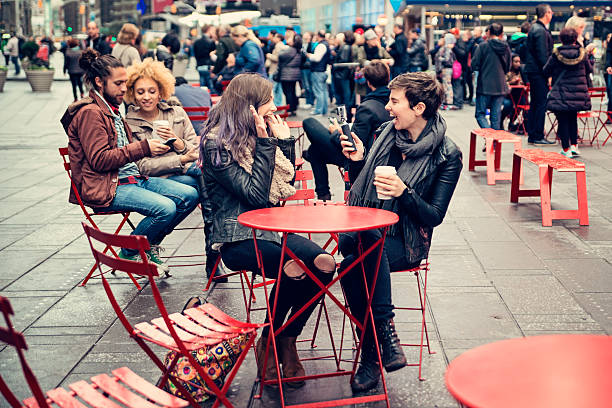 Girls in the foreground are playing with a small camera, mixed race couple in the back are looking at their phone, crowd of people is in the background. This was taken at that public space with red chairs and tables in Times Square, New York. Full length outdoors shot with copy space.