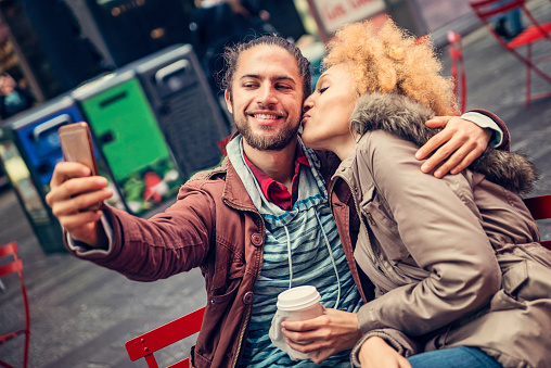 Tender moment between mixed race couple having coffee and doing a selfie in the city. Man has a beard and a ponytail, woman is mixed race with blond hair. Waist up horizontal shot on an overcast autumn day. This was taken at the public place with red chairs and tables in Times Square, New York.