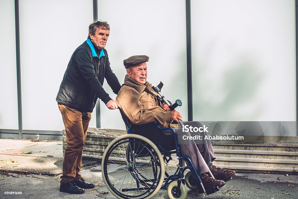 Senior Man in a Wheelchair with his Son, Europe Satisfied disabled senior man in the city with his male nurse, son, Slovenia, Europe. Sunny, cold, autumnal day.  Side view, copy space. Nikon D800, full frame, XXXL. Senior Men Stock Photo