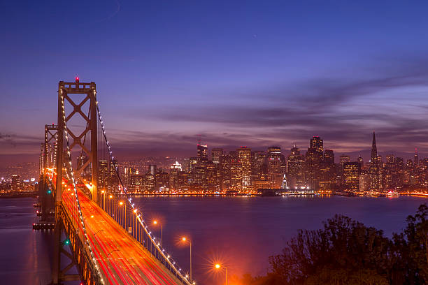 The San Francisco skyline stretches across the bay at night. Lights from the bridge and city illuminate the photo. Lights from the cars are blurred, creating movement. The city is reflected in the water below.