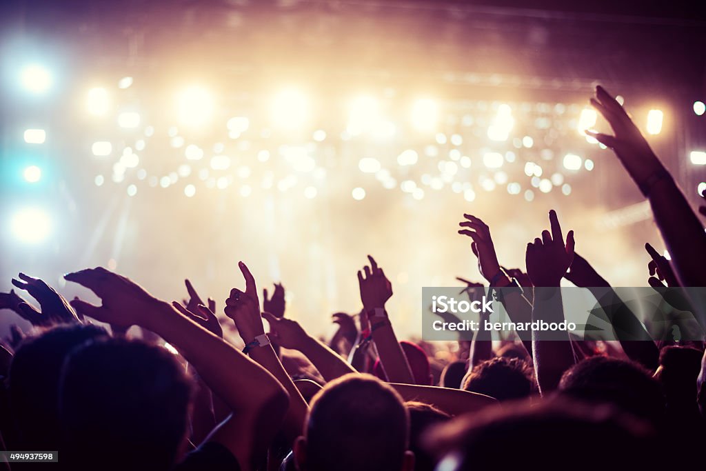 This party's on fire Audience with hands raised at a music festival and lights streaming down from above the stage. Soft focus, high ISO, grainy image. Music Festival Stock Photo