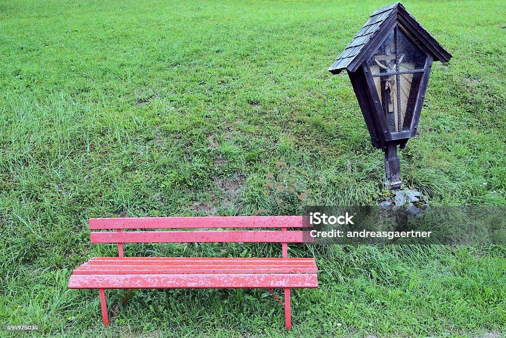 Pause for a moment A red bench on the edge of a meadow, near a wooden roadside calvary, weather-protected with a window glass. Photograph taken near Lunz, Austria. 2015 Stock Photo