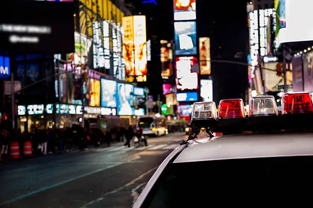 Times Square in New York City, USA at night.  Many billboards light up the streets.  Crowds of unrecognizable people walking across the street at a busy intersection in the bustling city.  View of the top of an NYPD police car in foreground.