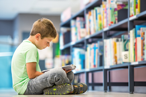 Schoolboy sitting on floor with book in library.