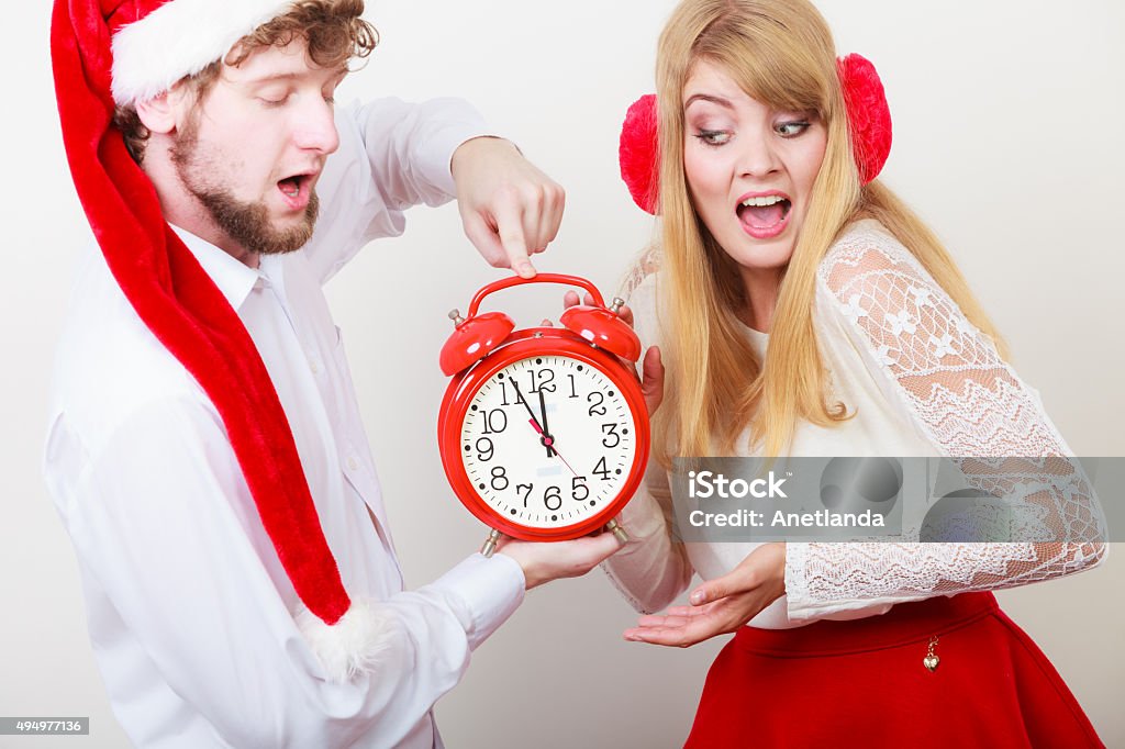 Scared couple woman and man with alarm clock. Scared cute couple holding alarm clock. Pretty woman in earmuffs and handsome man in santa claus hat on gray. Christmas time season concept. 12 O'Clock Stock Photo