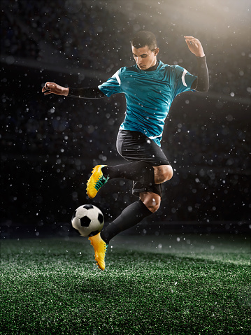 A male soccer player makes a dramatic play by jumping vertically. He attempts to kick the ball with his feet. The stadium is dark behind him. It's snowing and raining on the street.