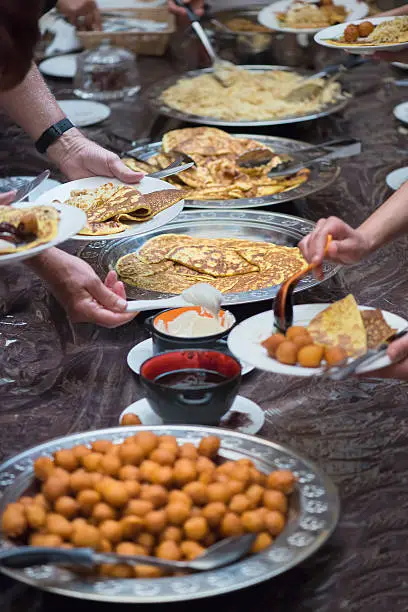 iStockalypse Dubai.  Personal perspective of a diner eating a traditional Emirati breakfast while guests serve themselves.   From front to back: dungaw (spiced chickpeas), date syrup, chabab (Emirati pancakes with cardamom and saffron), yogurt, balaleet (noodles with eggs, cardamom, saffron and raisins), ligamat (donuts).  Dishes are served on a plastic covered floor.  Dubai, UAE, Middle East, GCC.