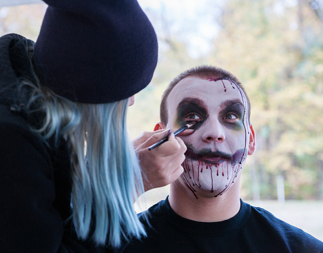 Indiana, USA - October 31, 2015: Woman wearing a blue wig applies stage makeup to a college aged male student in preparation for the 5K Zombie Run on Halloween