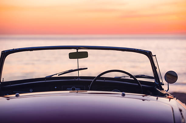 Convertible at the beach at sunset or sunrise. Car is parked at the edge of the sand looking at the view over the ocean. No people. Copy space. Rear view.